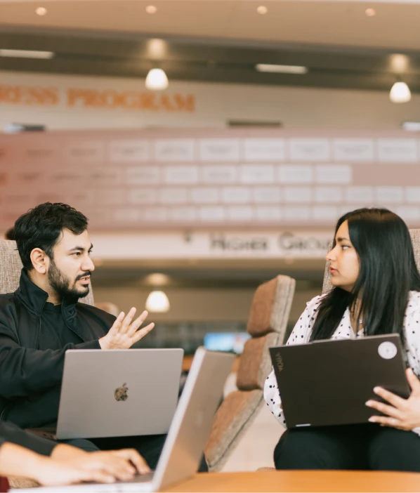 Two people talking across a lounge table, each with a laptop open
