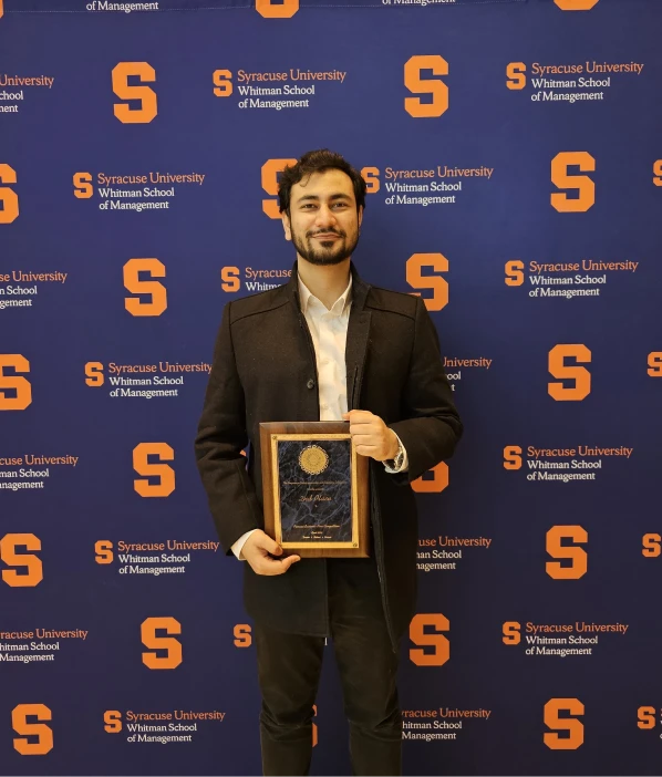 A man holding an award plaque in front of a Syracuse University Whitman School of Management backdrop