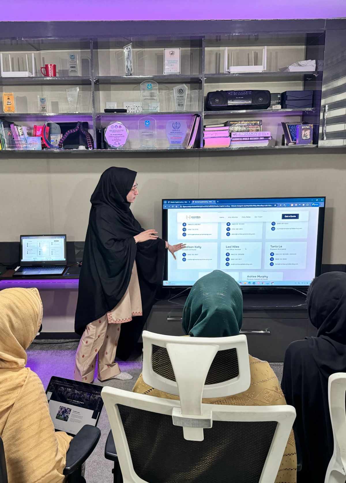 courses A woman in a black hijab and a tan outfit is standing in front of a screen, teaching a course to a group of students