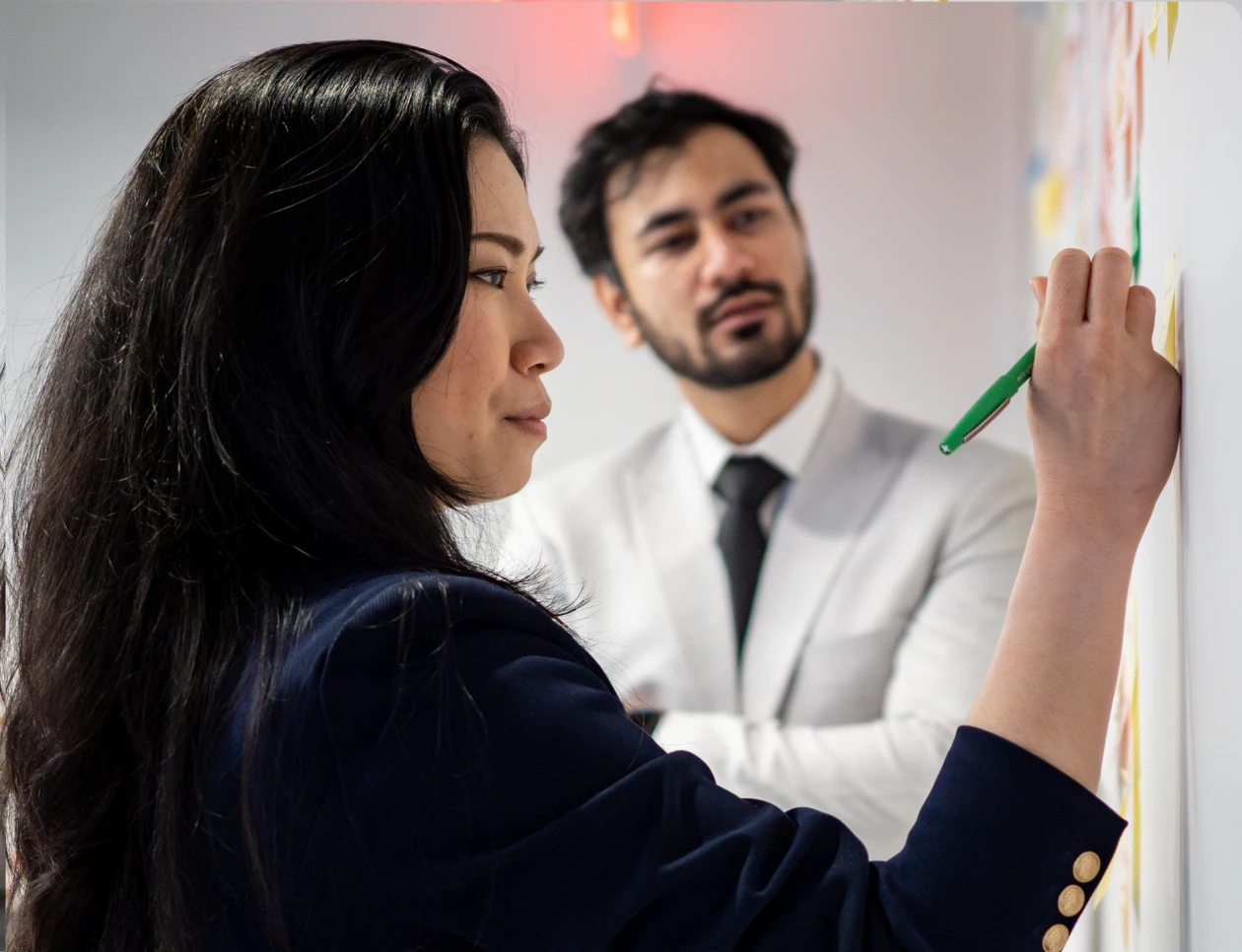 courses A woman with long dark hair is writing on a whiteboard with a green marker, while a man in a white shirt and black