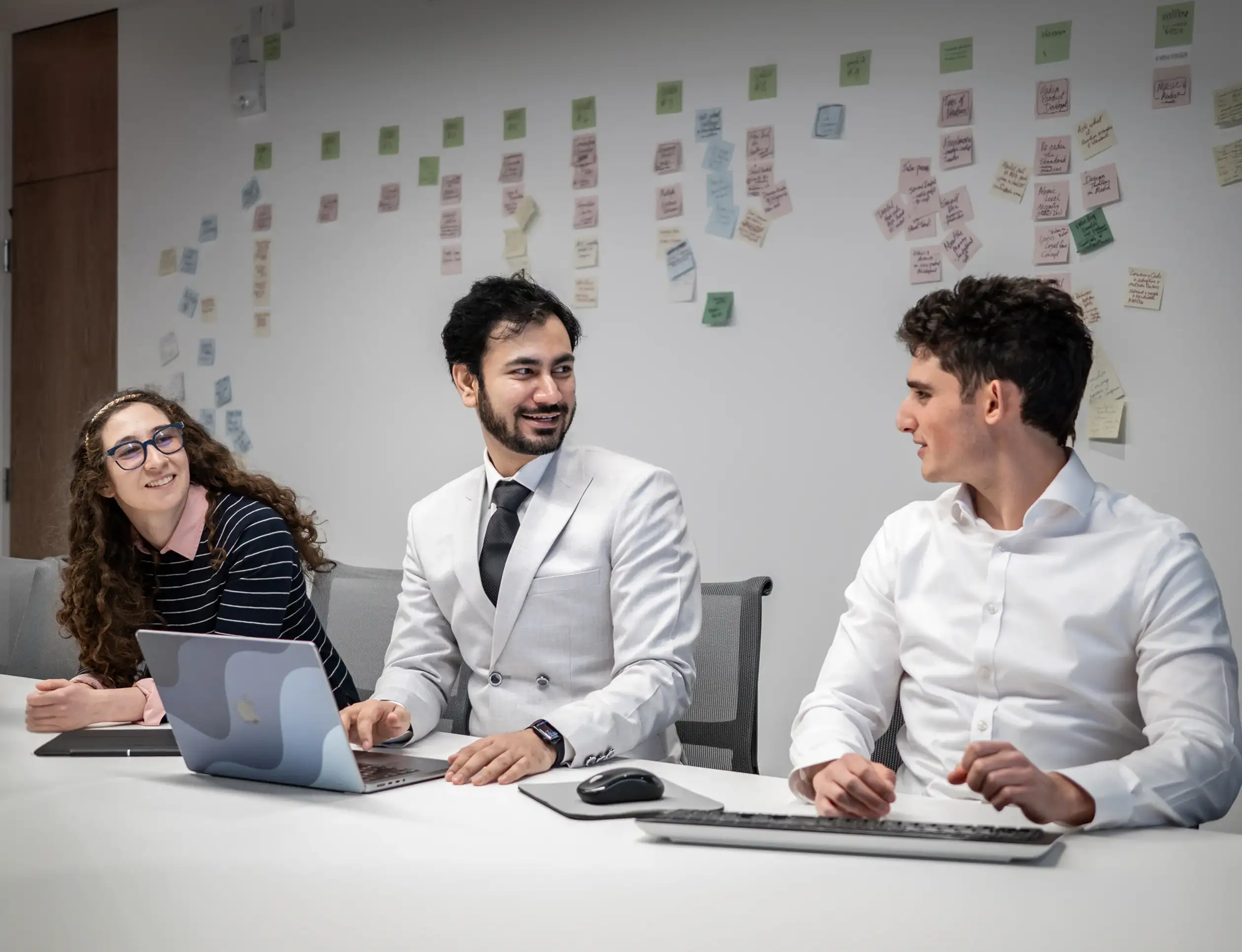 Three people talking at a desk with a laptop, in front of a wall covered in sticky notes
