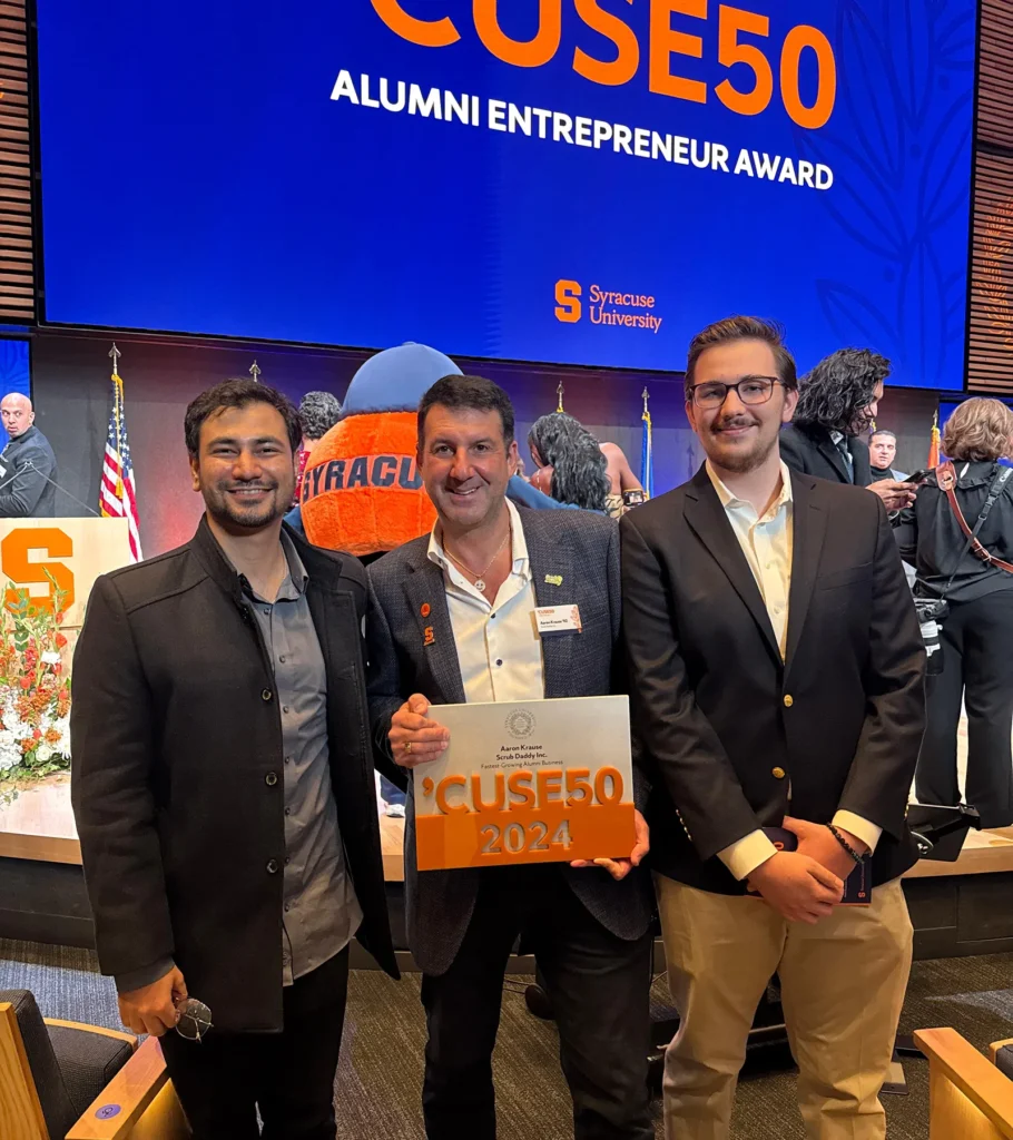 Waqar Hussain, Icon Pro Solutions CEO, with two other men holding a 'Cuse50 2024 award under a screen reading 'Cuse50 Alumni Entrepreneur Award, Syracuse University