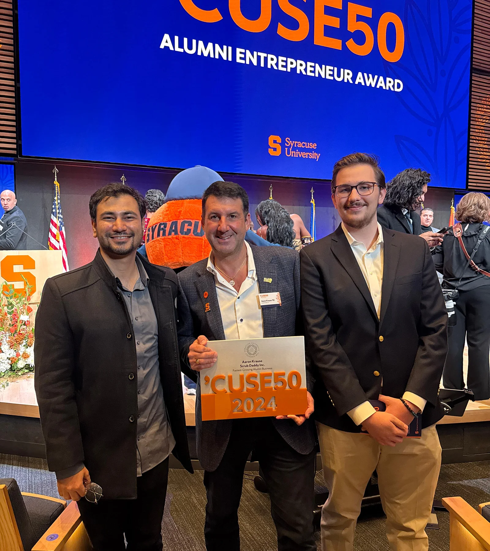 Waqar Hussain, Icon Pro Solutions CEO, with two other men holding a 'Cuse50 2024 award under a screen reading 'Cuse50 Alumni Entrepreneur Award, Syracuse University