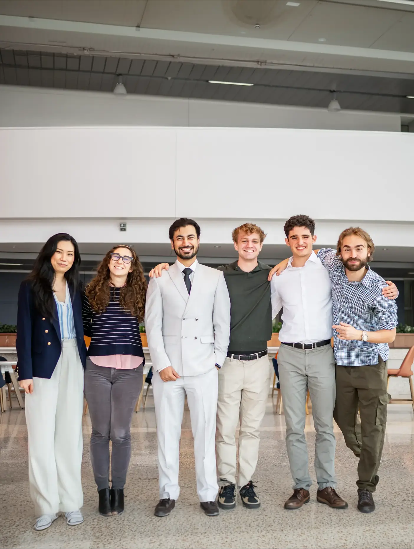 Six people standing arm in arm and smiling in a bright university hall